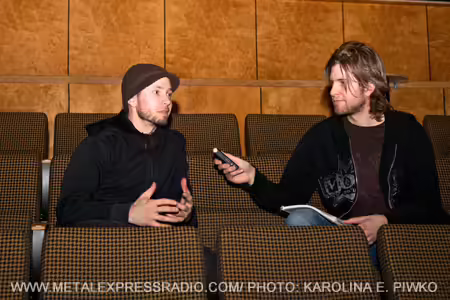 Photograph of two people seated in upholstered, patterned chairs, engaged in an interview. One person holds a handheld microphone towards the other. The image contains the text "WWW.METALEXPRESSRADIO.COM/PHOTO: KAROLINA E. PIWKO" at the bottom.