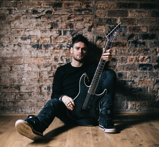 Fraser Edwards with his guitar in front of a brick wall