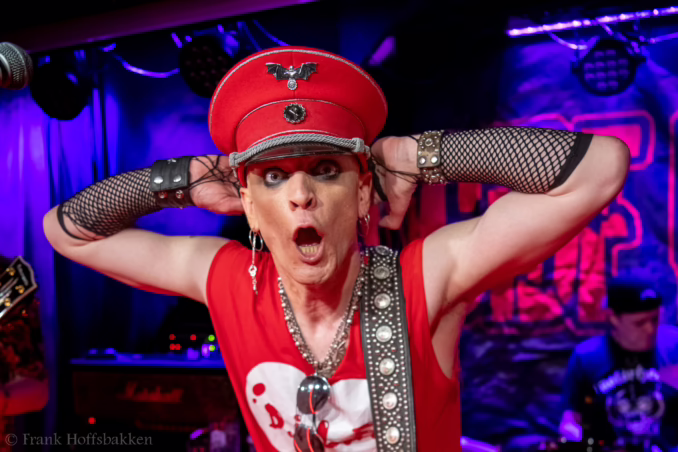A performer in a red military-style cap, fishnet sleeves, and studded accessories sings passionately on stage, illuminated by blue and purple stage lights.